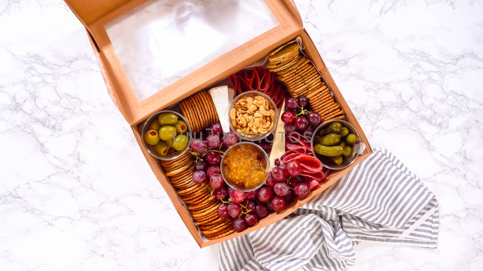 Flat lay. Hands are meticulously adding fresh red grapes to a bowl, complementing a beautifully arranged charcuterie box brimming with a variety of cheeses, olives, and cured meats, set against a sleek marble surface.