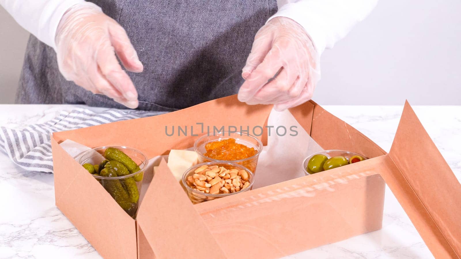 Hands are meticulously adding fresh red grapes to a bowl, complementing a beautifully arranged charcuterie box brimming with a variety of cheeses, olives, and cured meats, set against a sleek marble surface.