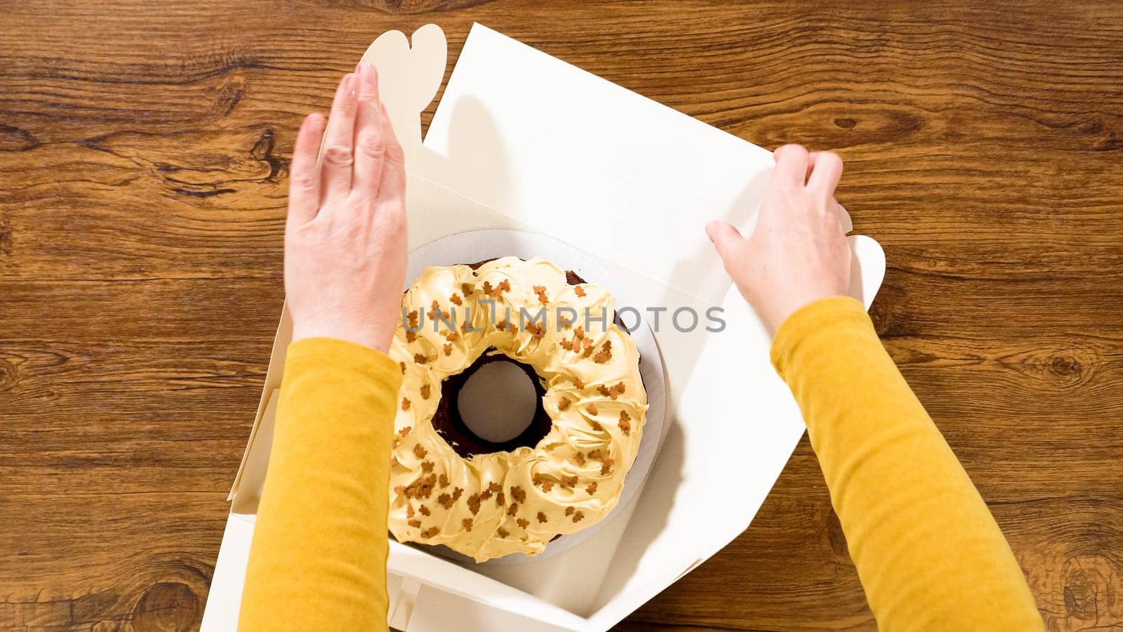 Flat lay. Carefully placing the gingerbread bundt cake, adorned with salted caramel frosting and gingerbread sprinkles, into a white paper bundt cake box with a clear window for gifting.