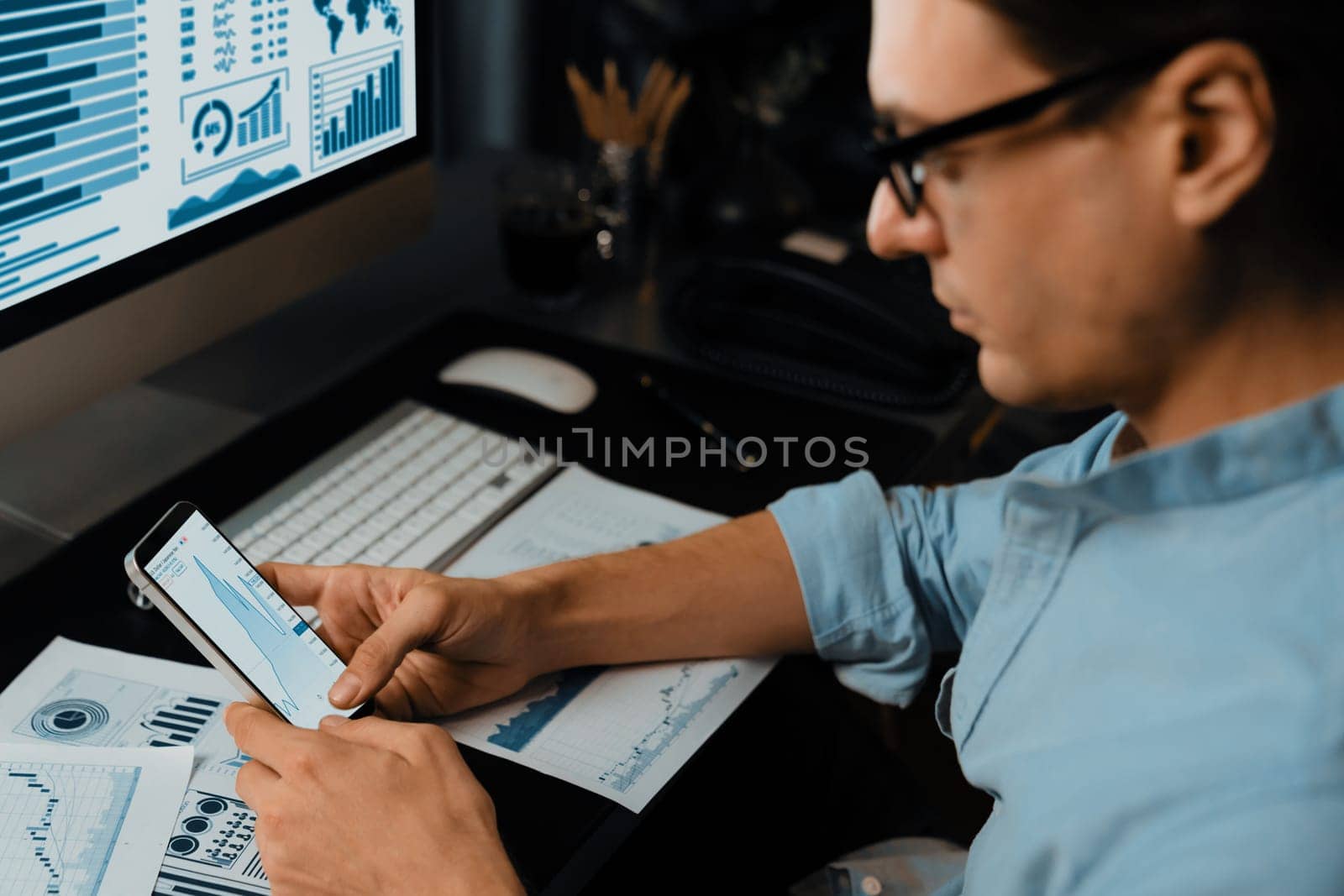 Smart businessman analyzing business paragraph dynamic database marketing analysis on smartphone surrounded paper sheet and pc monitor with company meeting report at home office at night. Pecuniary.
