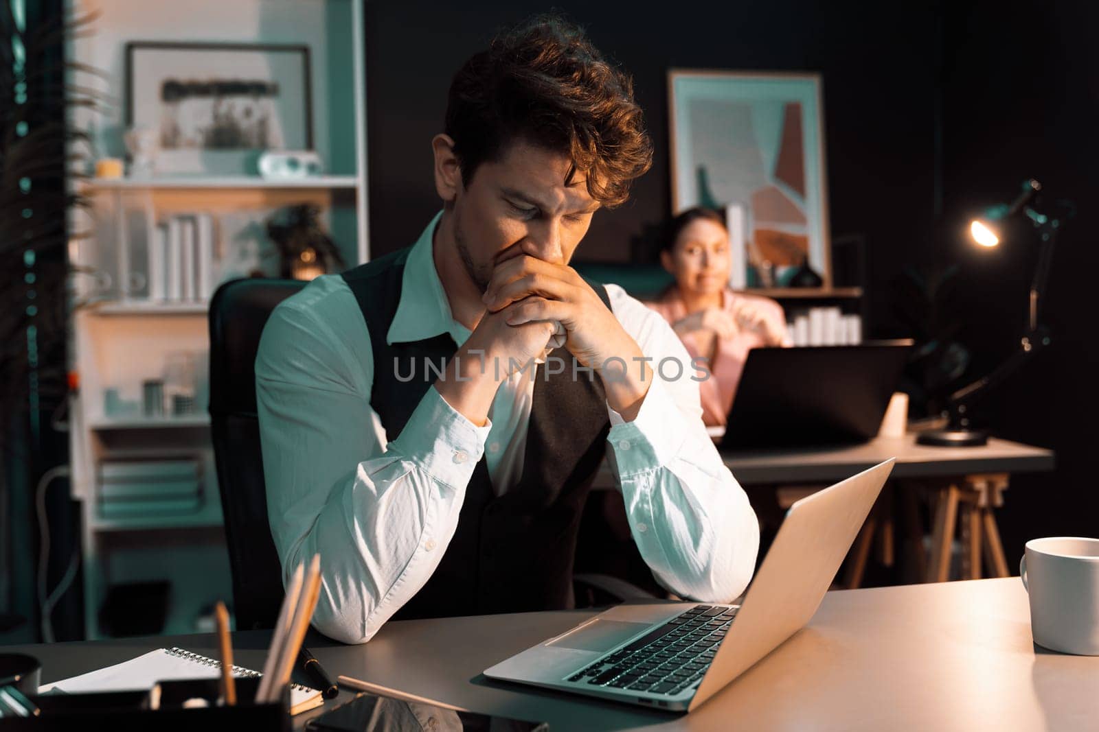 Sitting man with serious face thinking creative business project planning at night lighting time at modern office with blurry secretary checking paper report with folder background concept. Postulate.