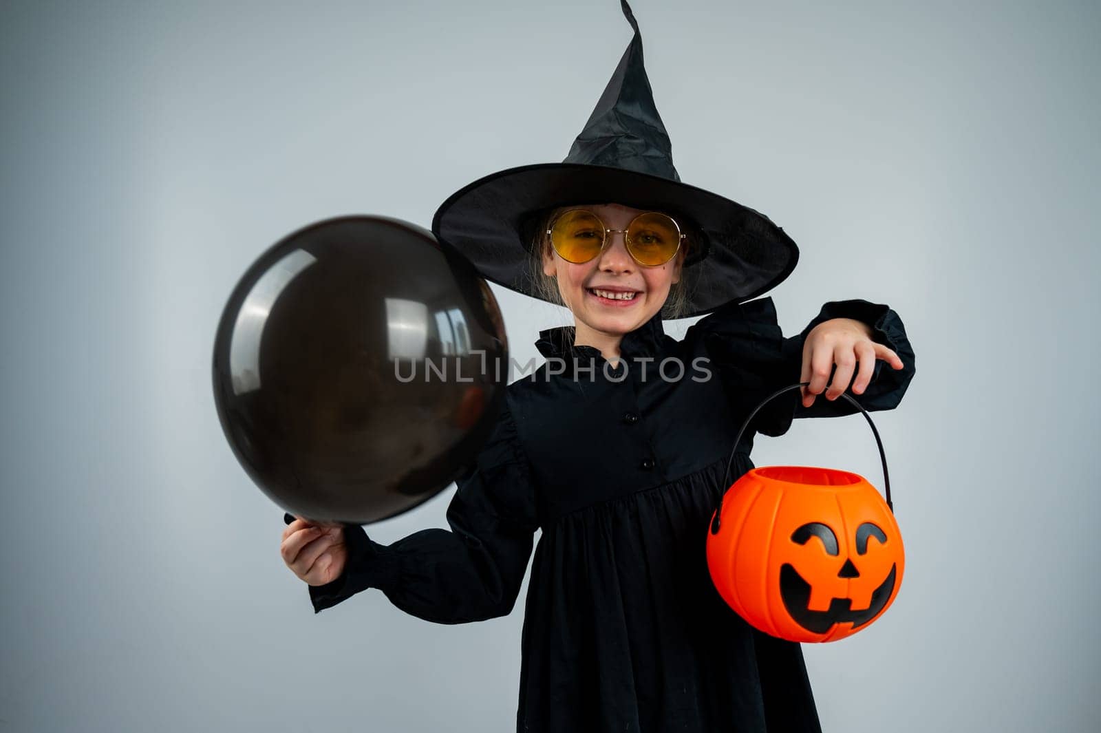 Portrait of a little Caucasian girl in a witch costume holding a cauldron and a black balloon on a white background
