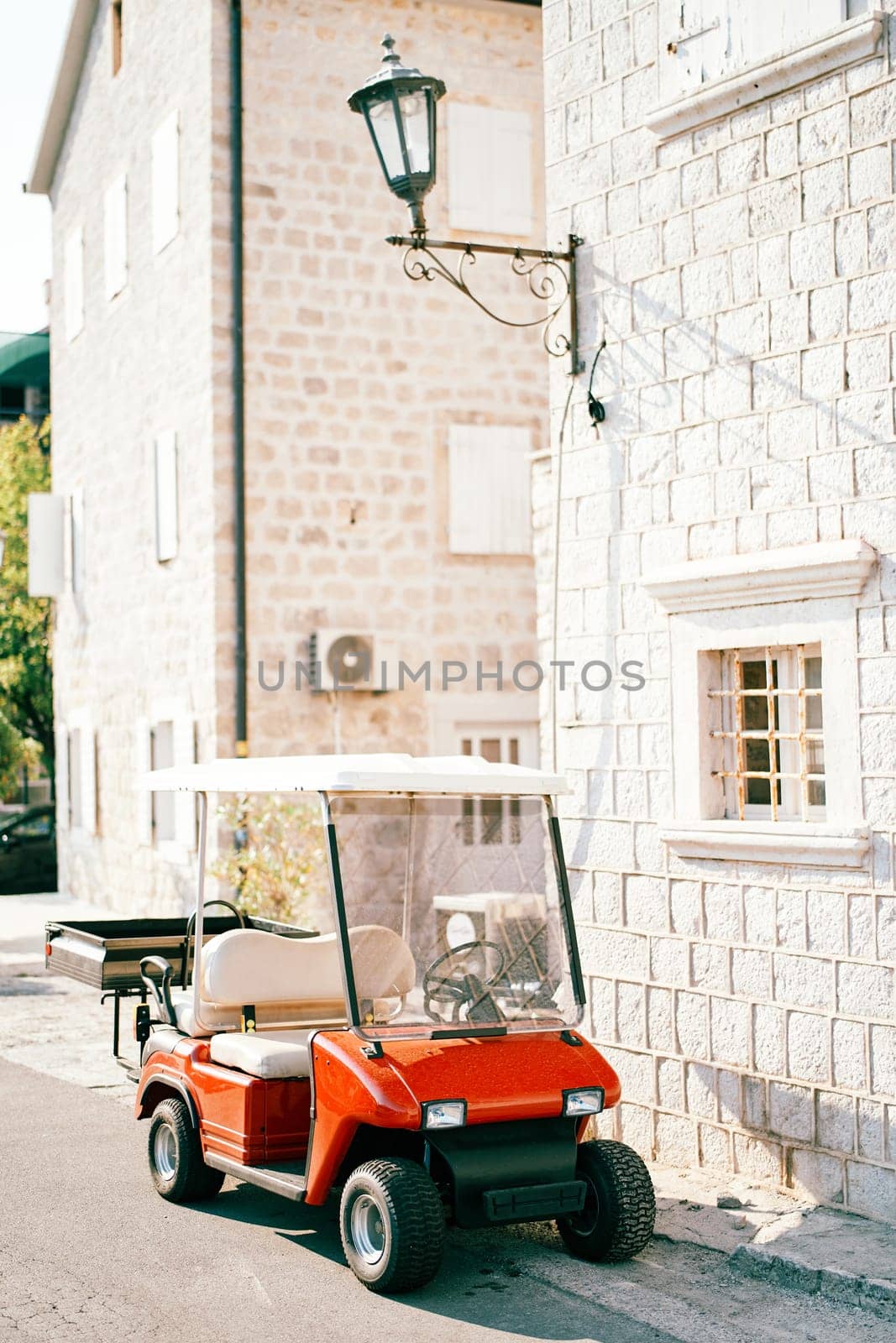 Red golf car with a trailer stands near an old stone house on the street. High quality photo