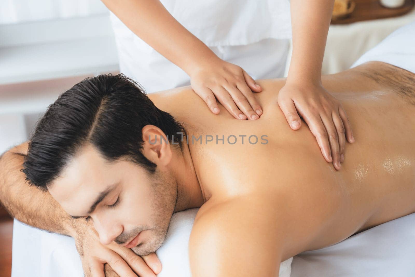 Caucasian man customer enjoying relaxing anti-stress spa massage and pampering with beauty skin recreation leisure in day light ambient salon spa at luxury resort or hotel. Quiescent