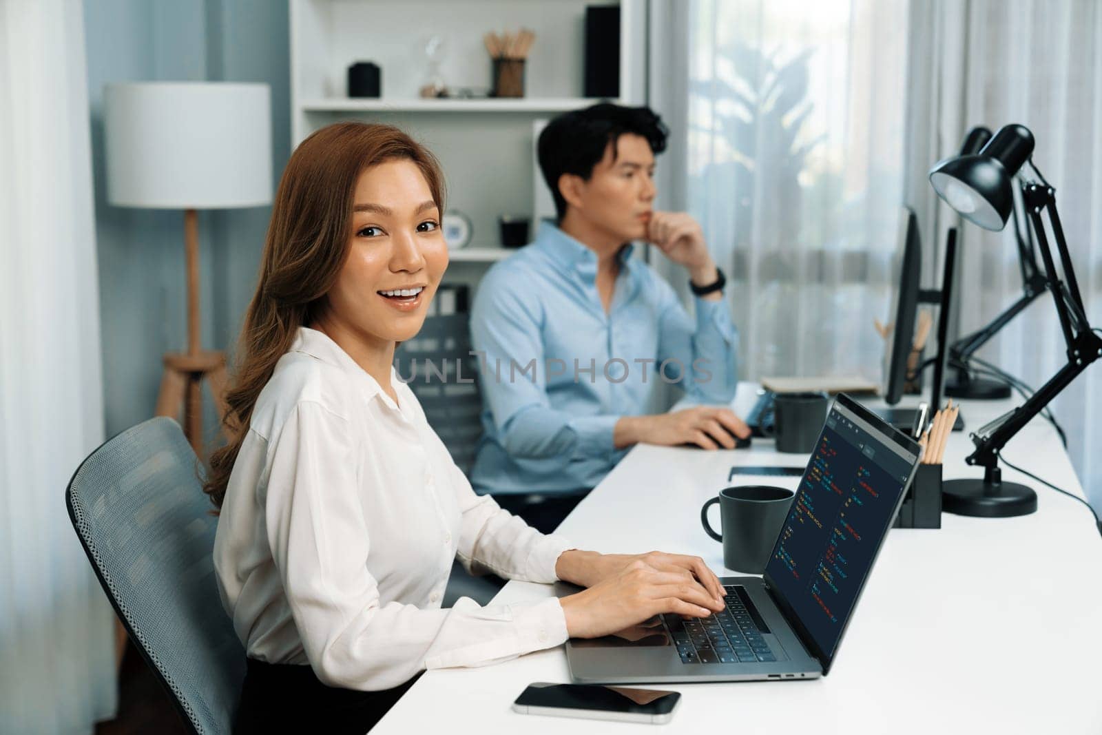 Profile smiling beautiful Asian businesswoman looking camera while typing on laptop at modern office on working desk casual day. Blurry background man colleague analyzing data market on pc. Infobahn.