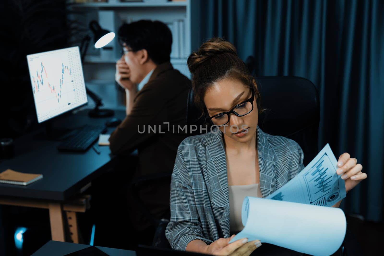 Businesswoman with stressful face looking document paperwork file while coworker trading stock market at back at over late night time at modern office, working on startup company project. Infobahn.