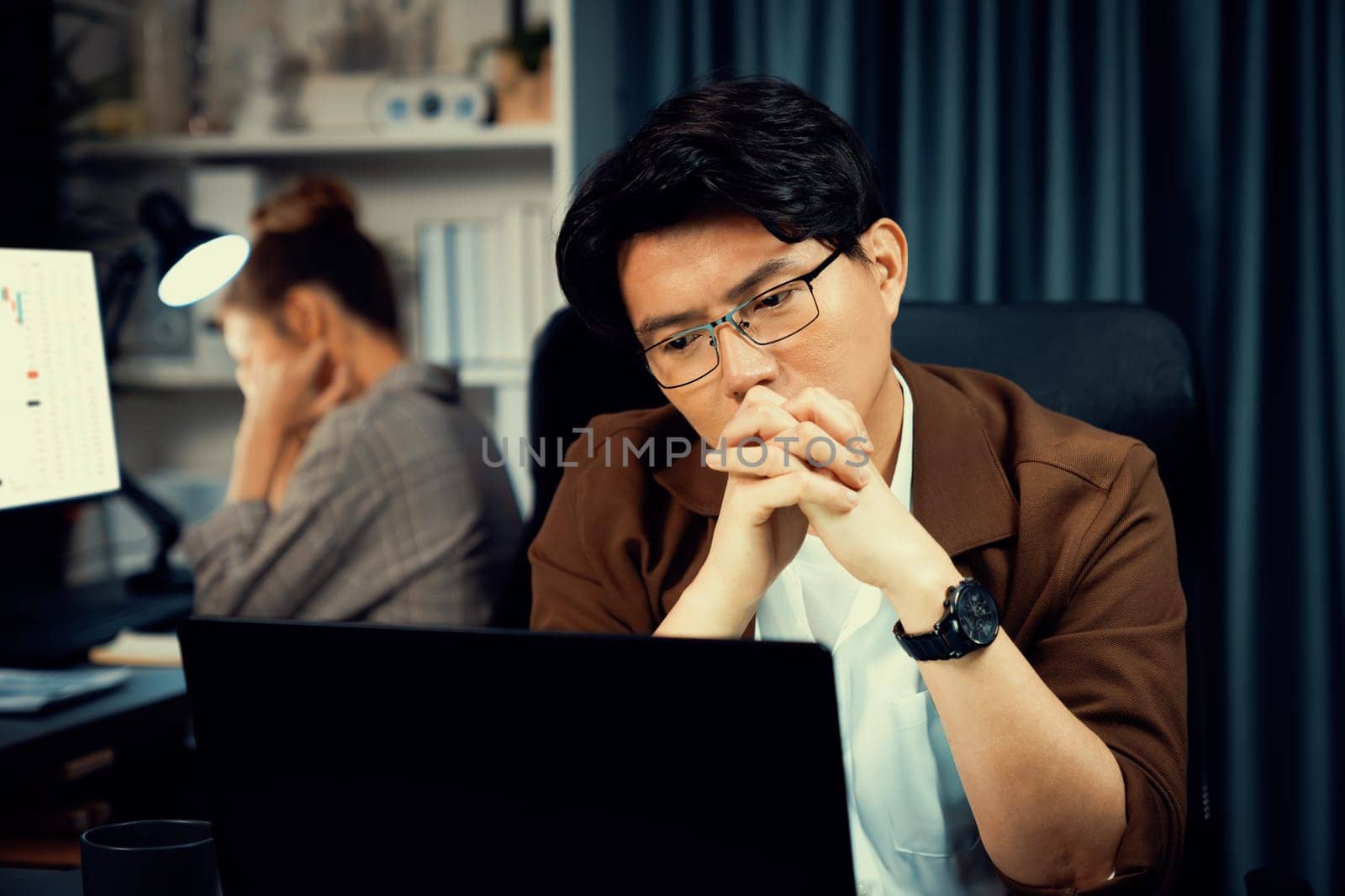 Businessman with stressful face working on laptop waiting email while coworker trading stock exchange investment at late night time at modern office, working on startup company project job. Infobahn.