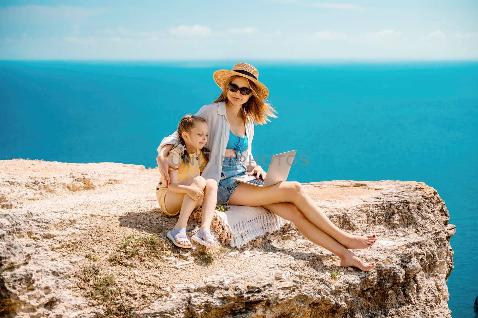 A woman and a child are sitting on a rock overlooking the ocean. The woman is using a laptop while the child looks on. Concept of relaxation and bonding between the two