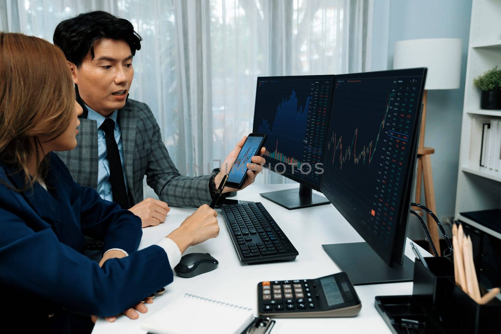 Smiling businessman showing dynamic stock market data on mobile phone to woman writing to memo for analyzing profit value currency rate online website program application at home office. Infobahn.