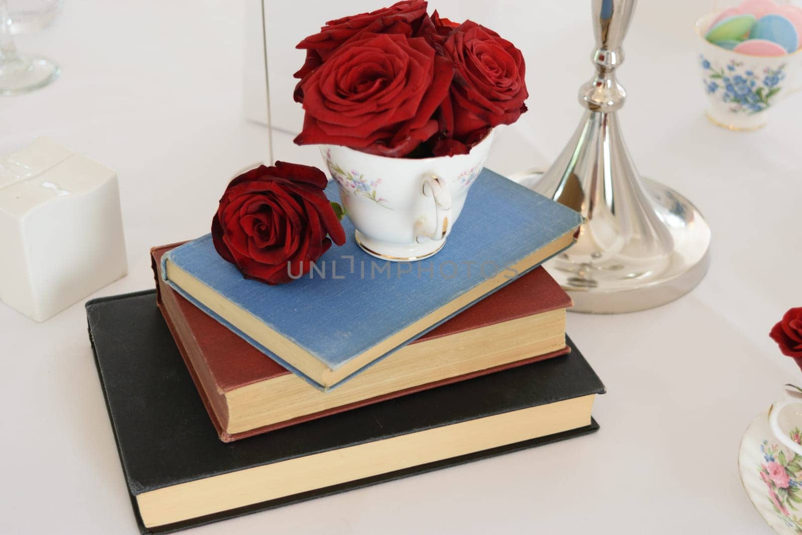 A stack of three books with a teacup filled with red roses on top, placed on a white tablecloth.