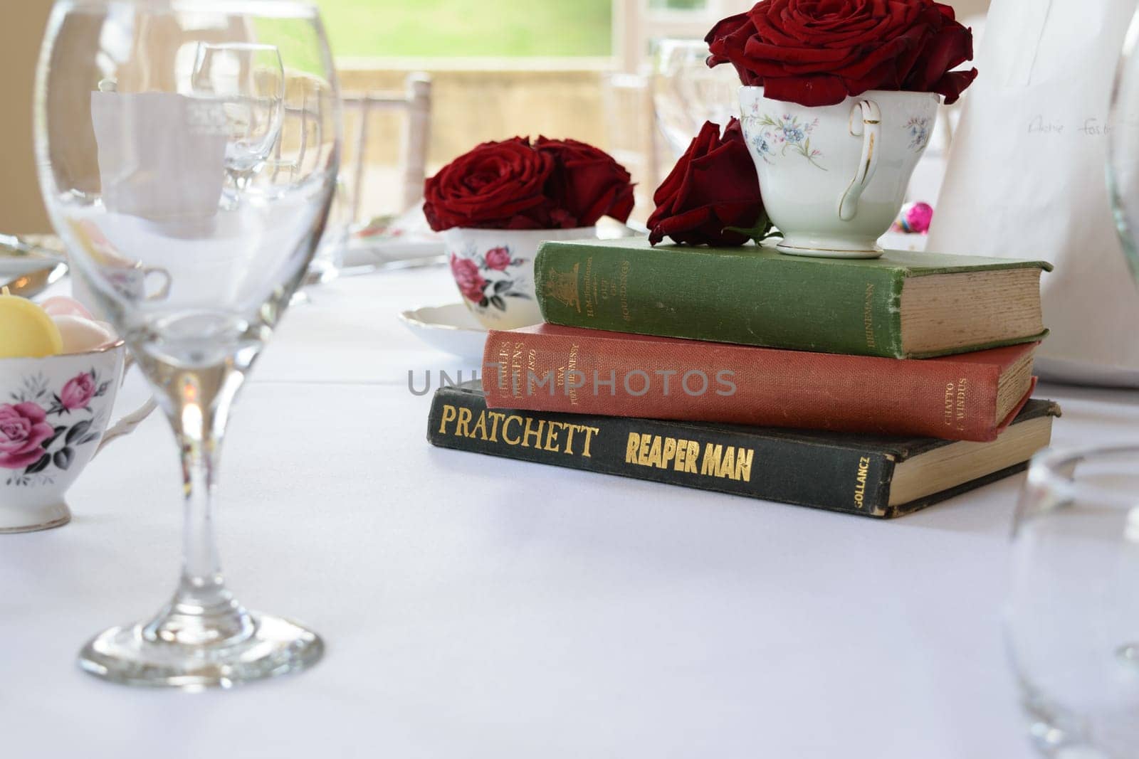A table setting with wine glasses, teacups with red roses, and a stack of books.