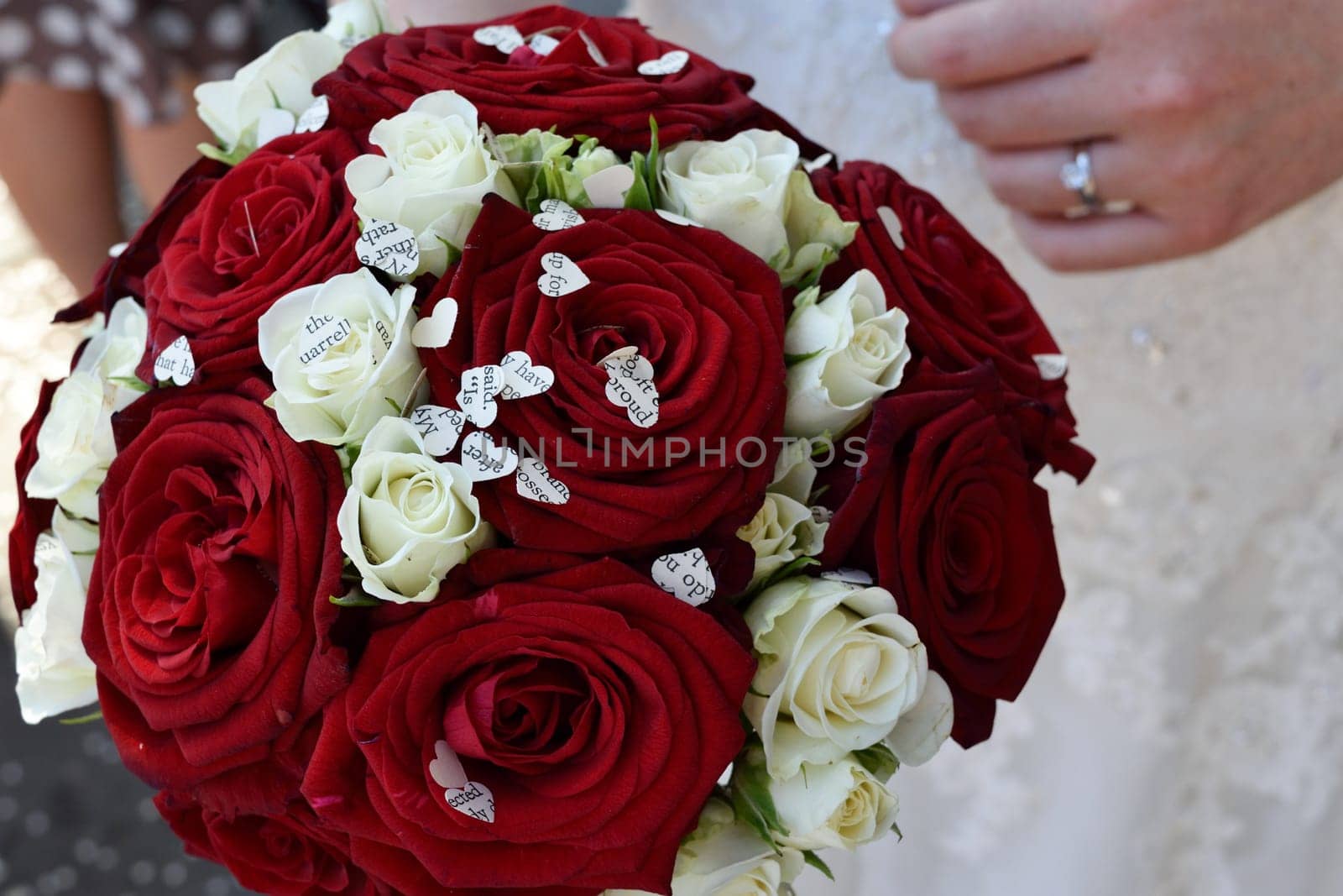 A close-up of a bridal bouquet featuring red and white roses with small heart-shaped paper cutouts scattered among the flowers. The bride's hand, wearing a wedding ring, is visible holding the bouquet.