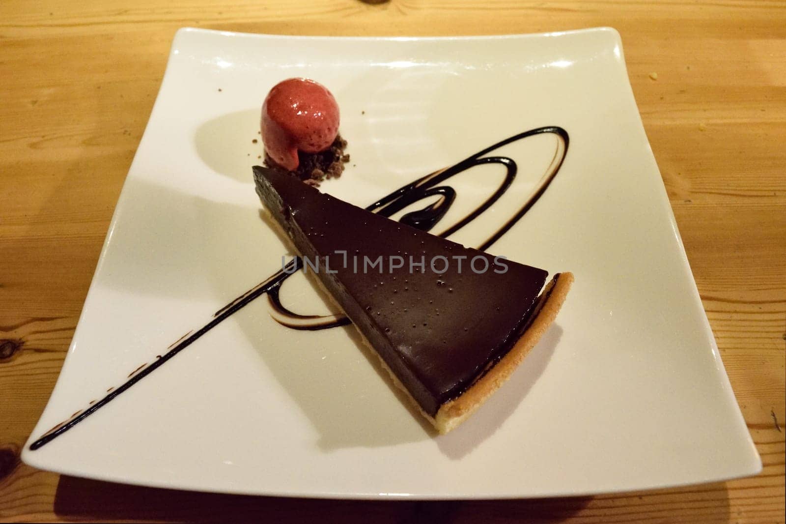 A slice of chocolate tart with a scoop of raspberry sorbet on a white plate, garnished with chocolate sauce, placed on a wooden table.