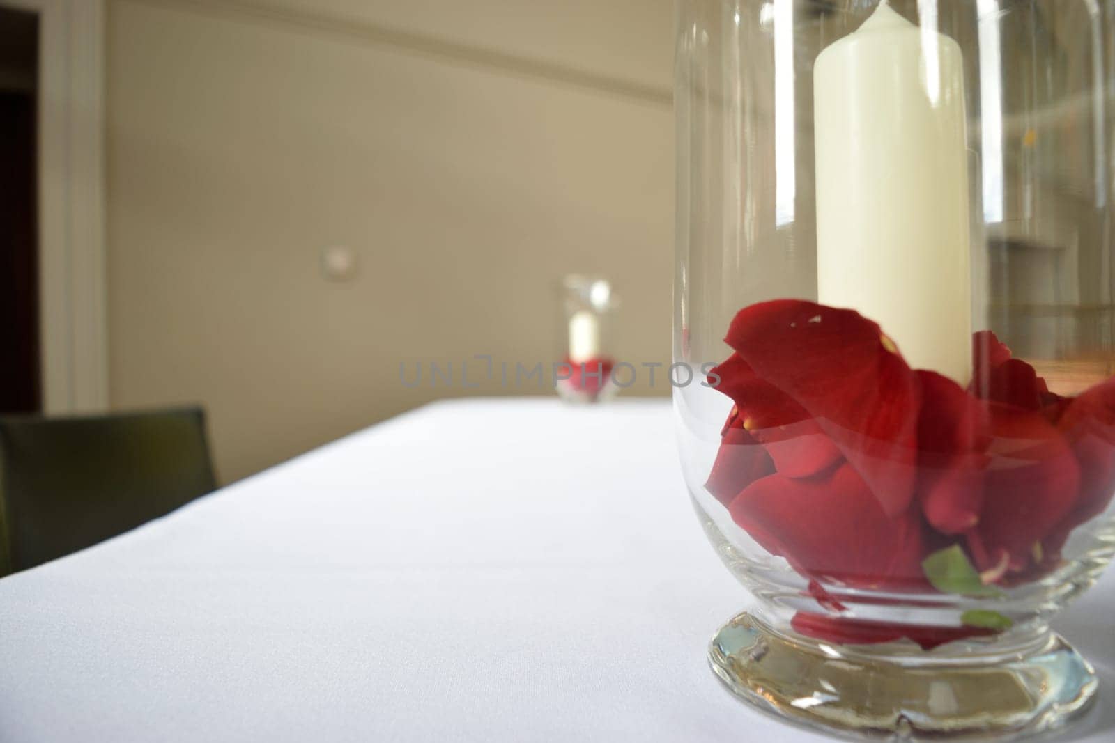 A white candle surrounded by red rose petals in a glass vase on a white tablecloth, with another similar arrangement blurred in the background.