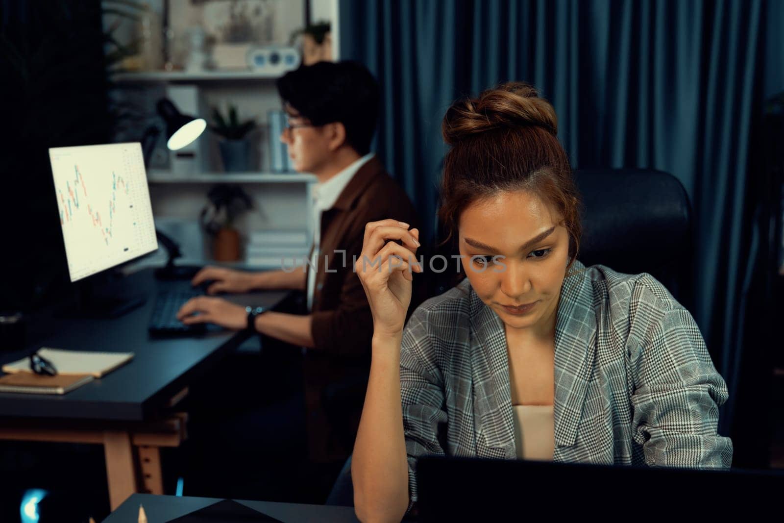 Serious face of thinking businesswoman focusing computer to report data analysis social media online project job with coworker working at back side at over late night time at modern office. Infobahn.