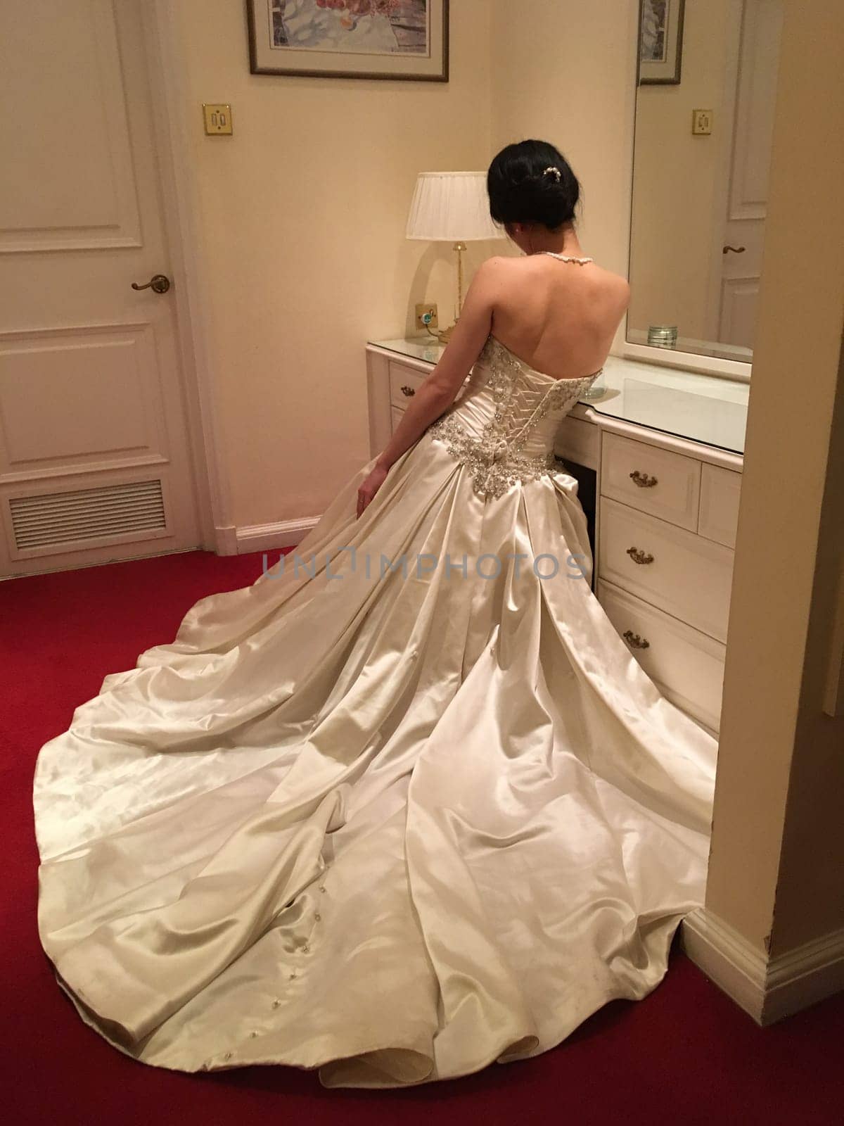 A bride in a white wedding dress with a long train sits on a vanity table in a room with red carpet and white walls.