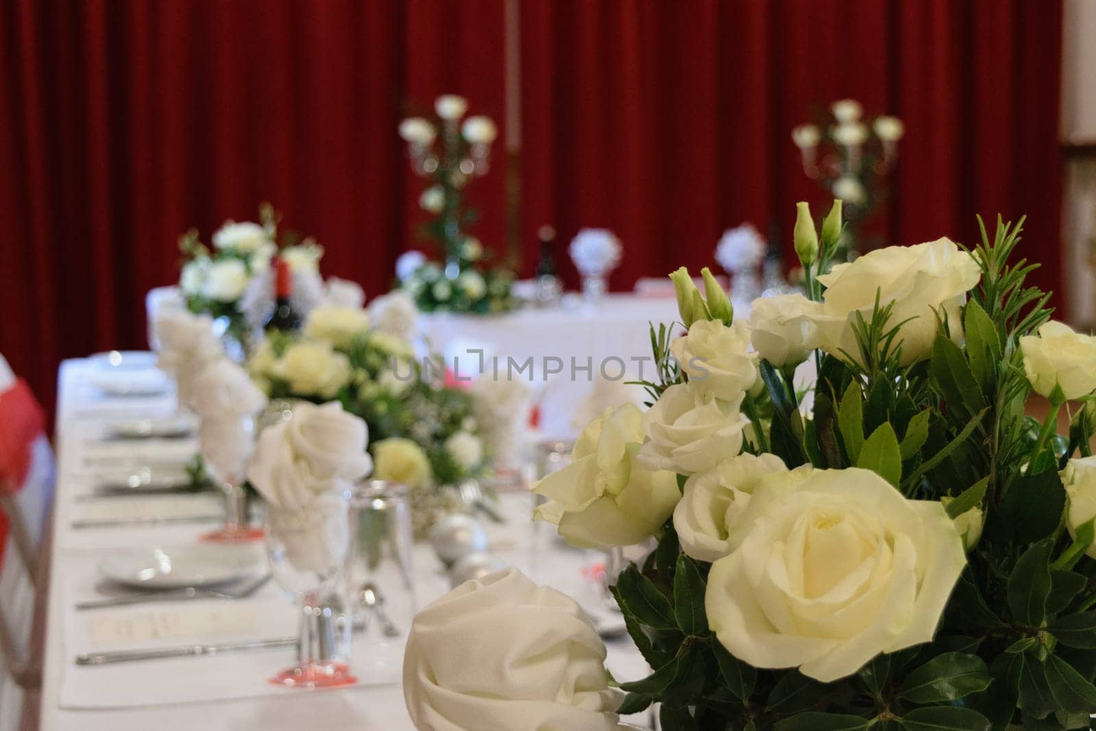 A beautifully decorated wedding reception table with white roses and elegant table settings. The background features red curtains and additional floral arrangements.