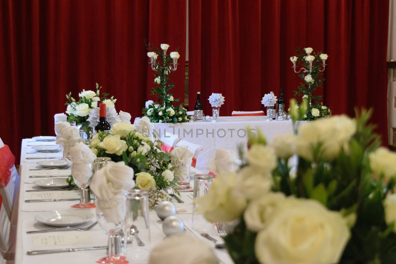 Elegant wedding reception setup with white floral arrangements, red curtains, and a long dining table.