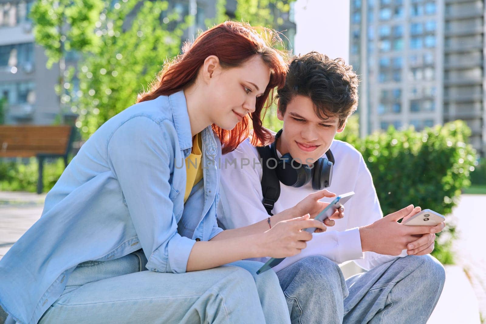 Teenage youth friends guy and girl university college students sitting outdoor on campus steps talking laughing using smartphone. Technology, lifestyle concept