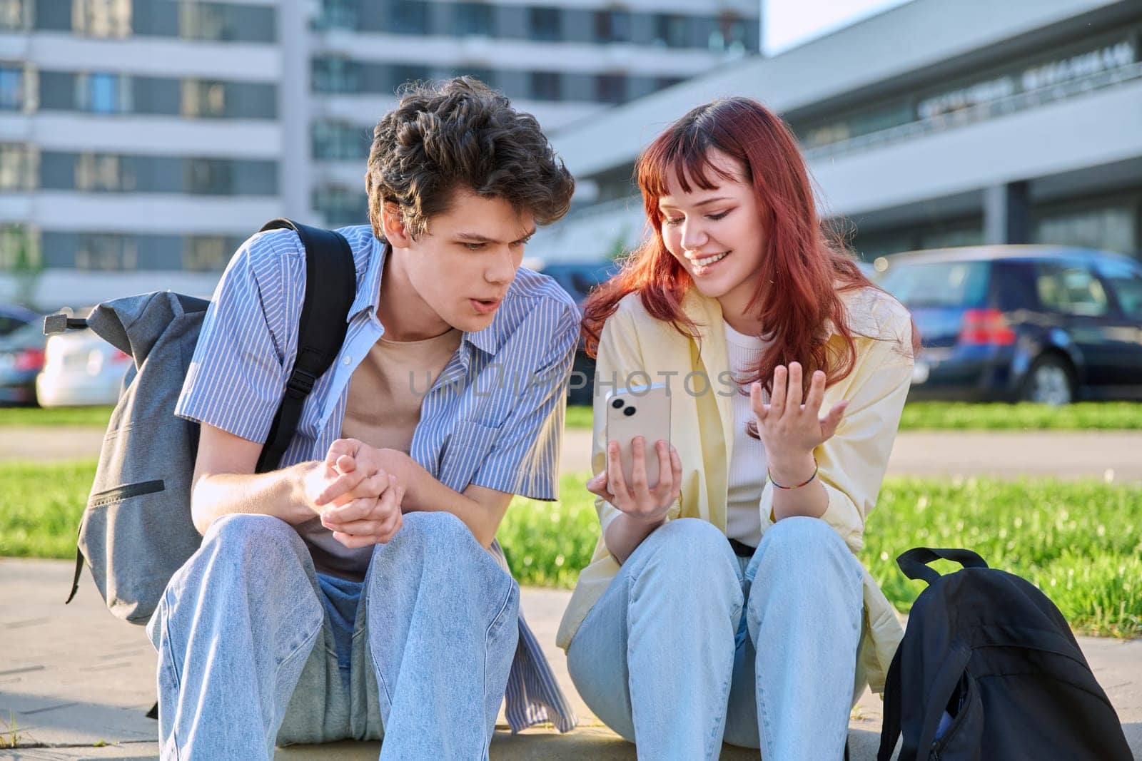 Friends young guy and girl university college students talking together, looking at smartphone outdoor, city modern buildings background. Lifestyle, communication, youth 19-20 years old, urban style