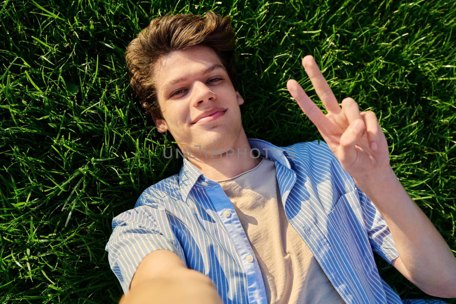 Top view, happy handsome smiling young male looking at camera, lying on background of green lawn grass, showing hand gesture peace victory success