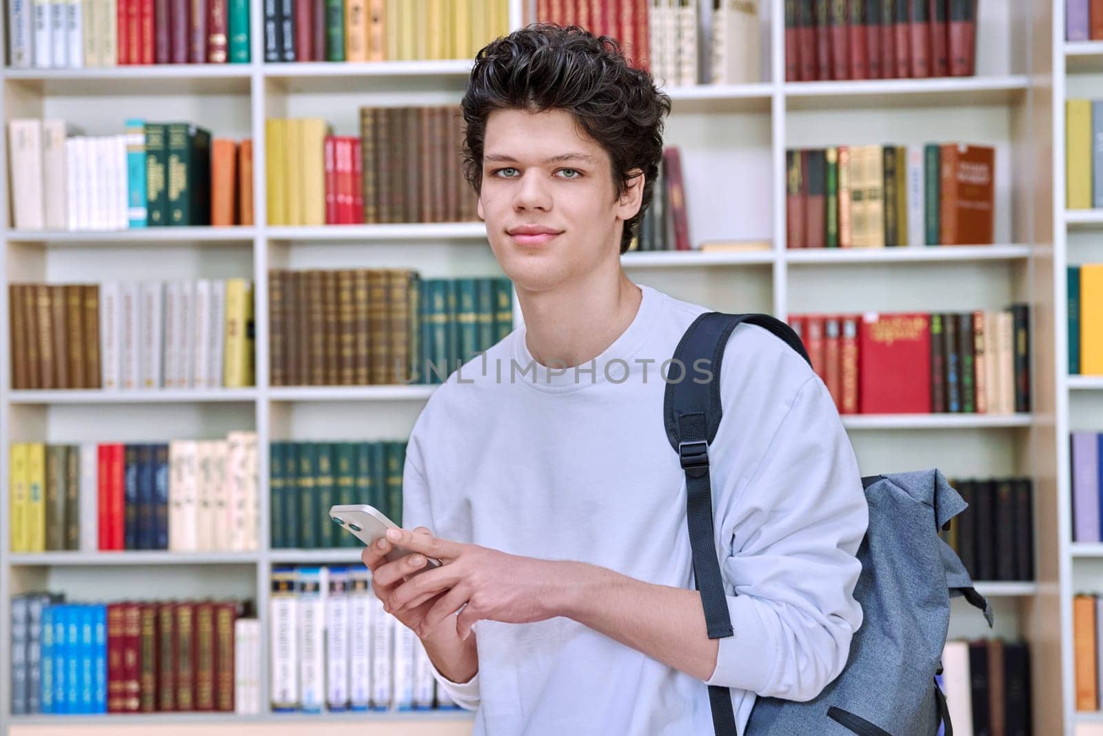 Teenage male student with backpack using smartphone, inside college building, in library. Technologies, mobile educational apps applications, services, e-learning concept