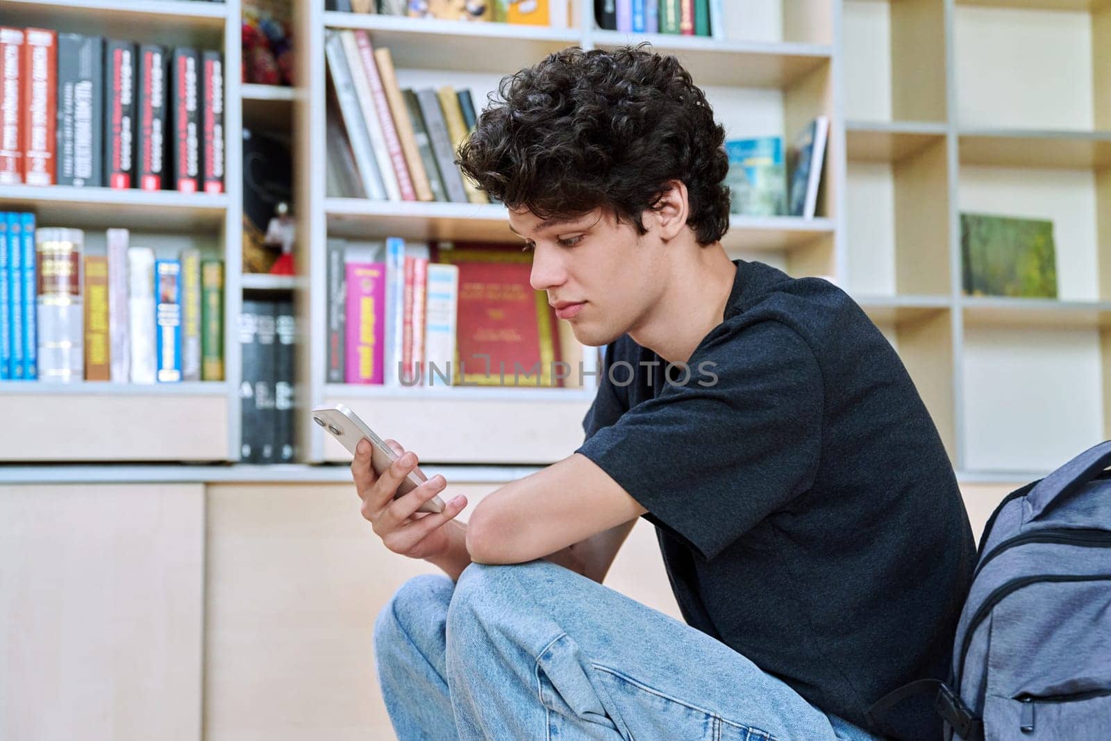 Relaxed handsome young male college student using smartphone, sitting inside library classroom. Teenager guy 19-20 years old, education, internet technology, mobile applications, lifestyle, youth