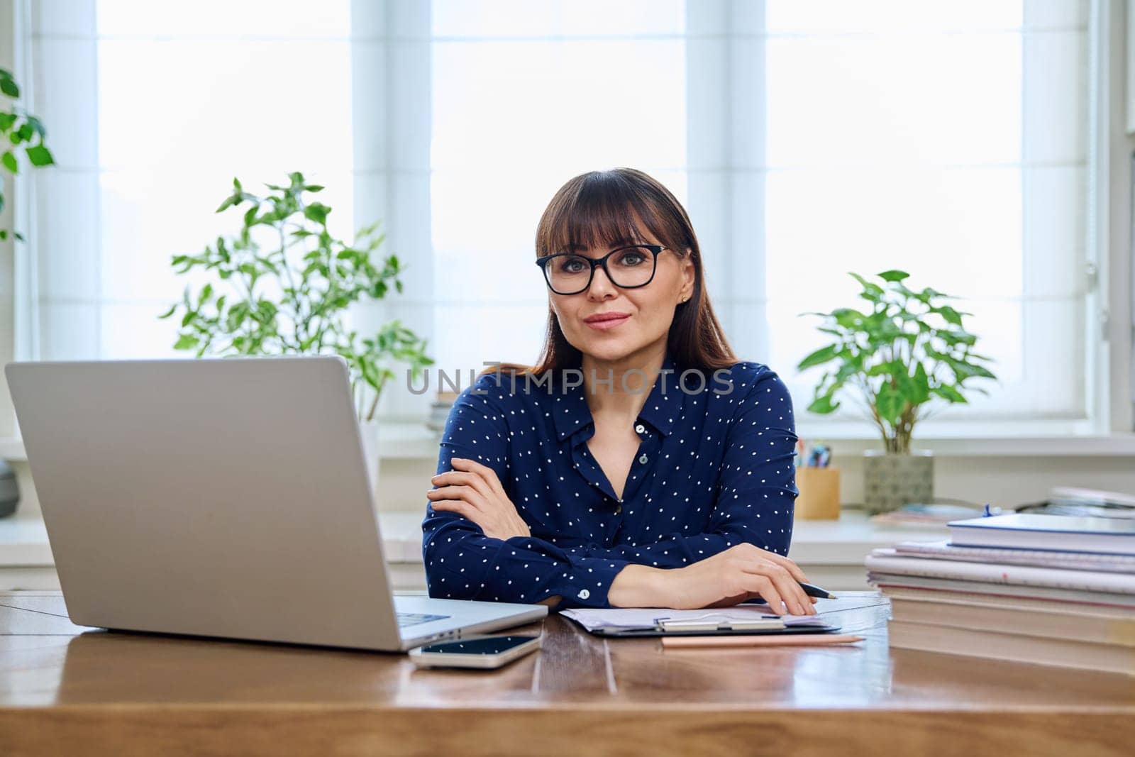 Middle-aged smiling woman working with laptop computer business papers, sitting at desk in home office interior, looking at camera. Work, remote business, freelance, technology, 40s people concept