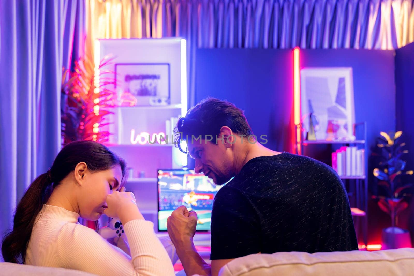 Man winner fighting gamer on video game beside woman loser challenge at back side view. Couple joyful playing on TV using joysticks in studio room in red blue neon light bulb at comfy home. Postulate.