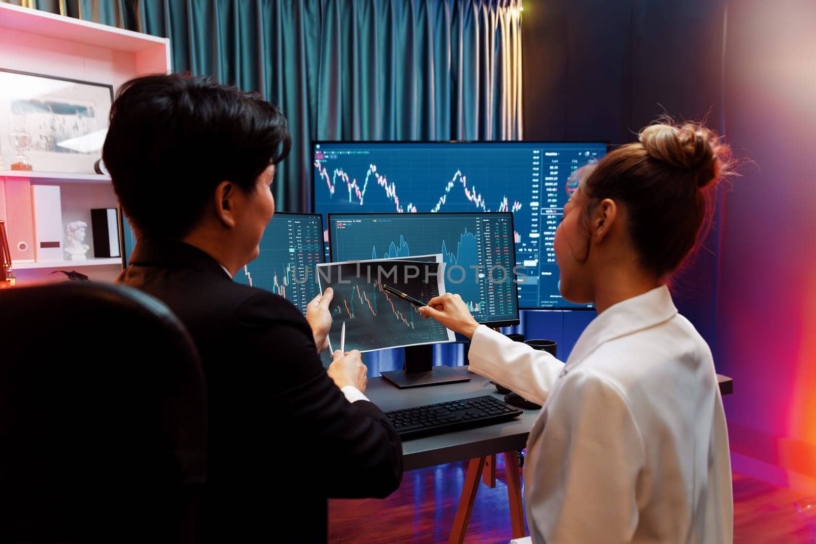 Asian businessman showing dynamic stock exchange graph paper with partner to discuss profit value online investment on website program application, analyzing report at neon light at night. Infobahn.