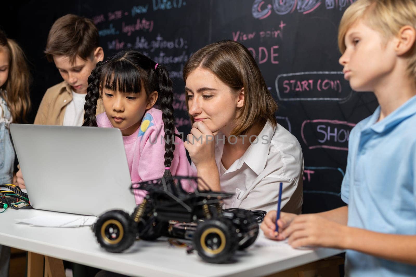 Skilled kids learning to program remote car. Schoolgirl in pink cloth use laptop for coding. Other child watch her code while teacher watch her. motherboard and electric wire also on table. Erudition.