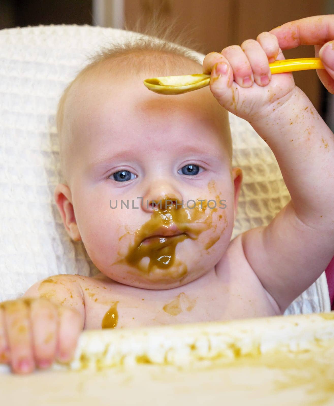 A cute baby with blue eyes has broccoli puree smeared all over their face. It looks like the baby is enjoying their meal while sitting at home. by Busker