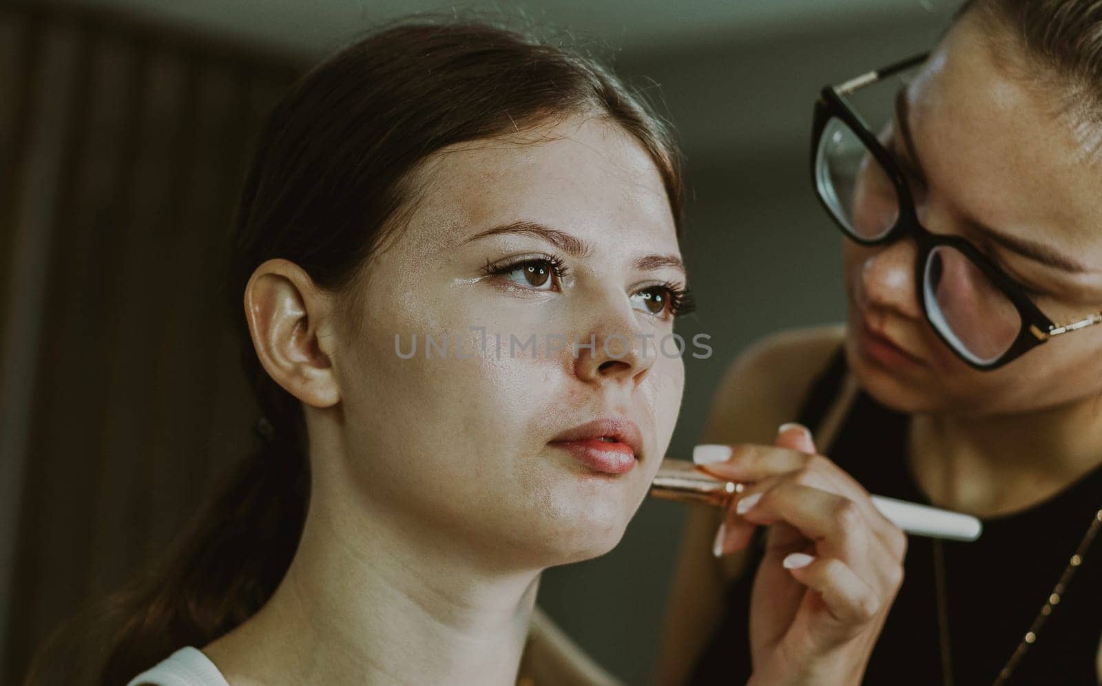 One young handsome Caucasian makeup artist applies blush with a brush to the cheekbones of a girl sitting in a chair early in the morning in a beauty salon, close-up side view. Step by step.