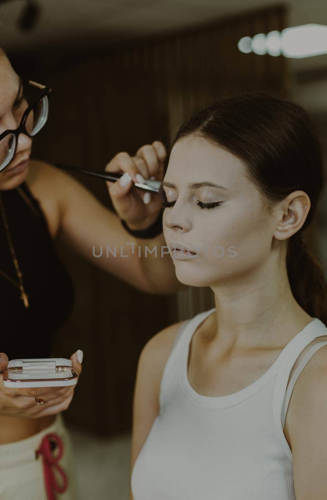 One young handsome Caucasian makeup artist applies foundation powder with a fluffy brush to the temples of a girl sitting in a chair early in the morning in a beauty salon, close-up side view. Step by step.