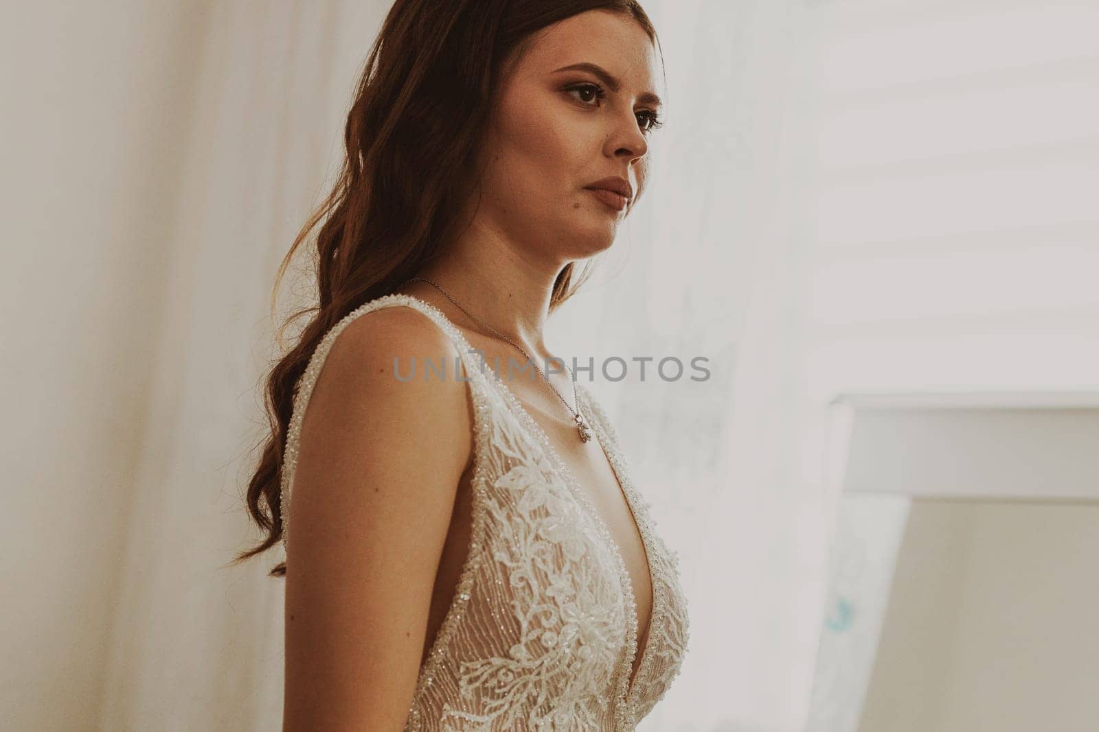 Portrait of a beautiful brunette bride in a white wedding dress standing in a room looking at herself in a mirror.