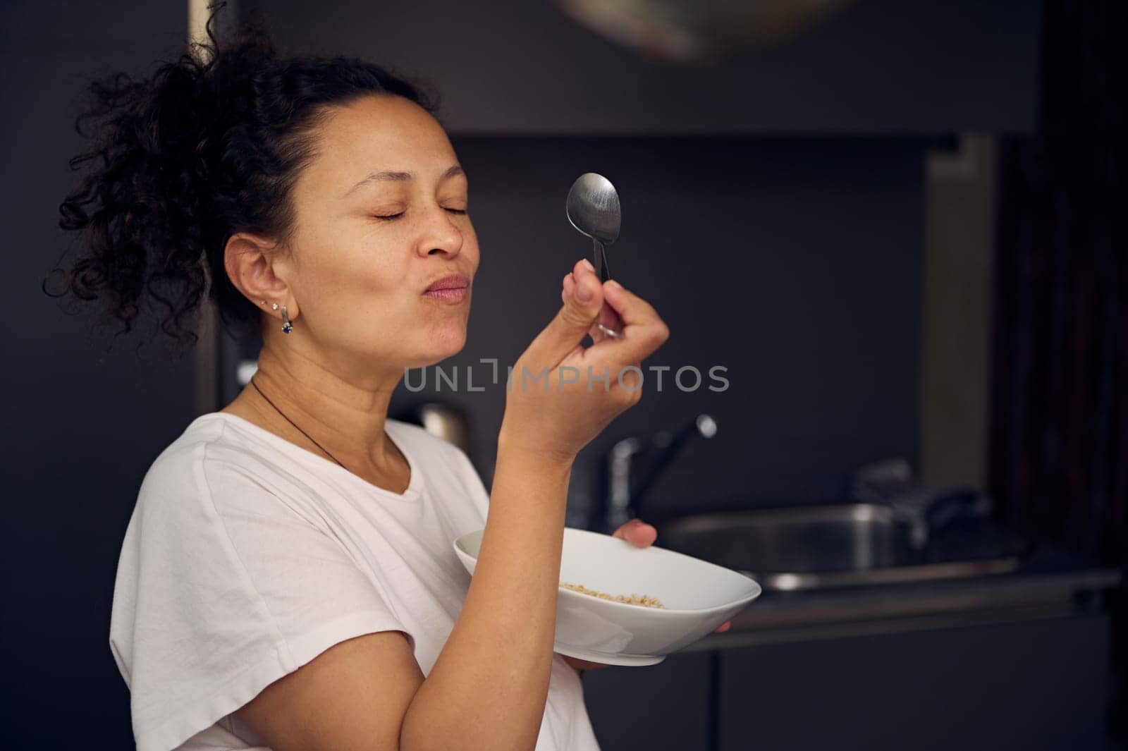 Authentic portrait of happy young woman enjoying her healthy breakfast in the morning. Smiling multi ethnic female in white pajamas, holding a muesli bowl and a spoon, eating oat flakes for breakfast