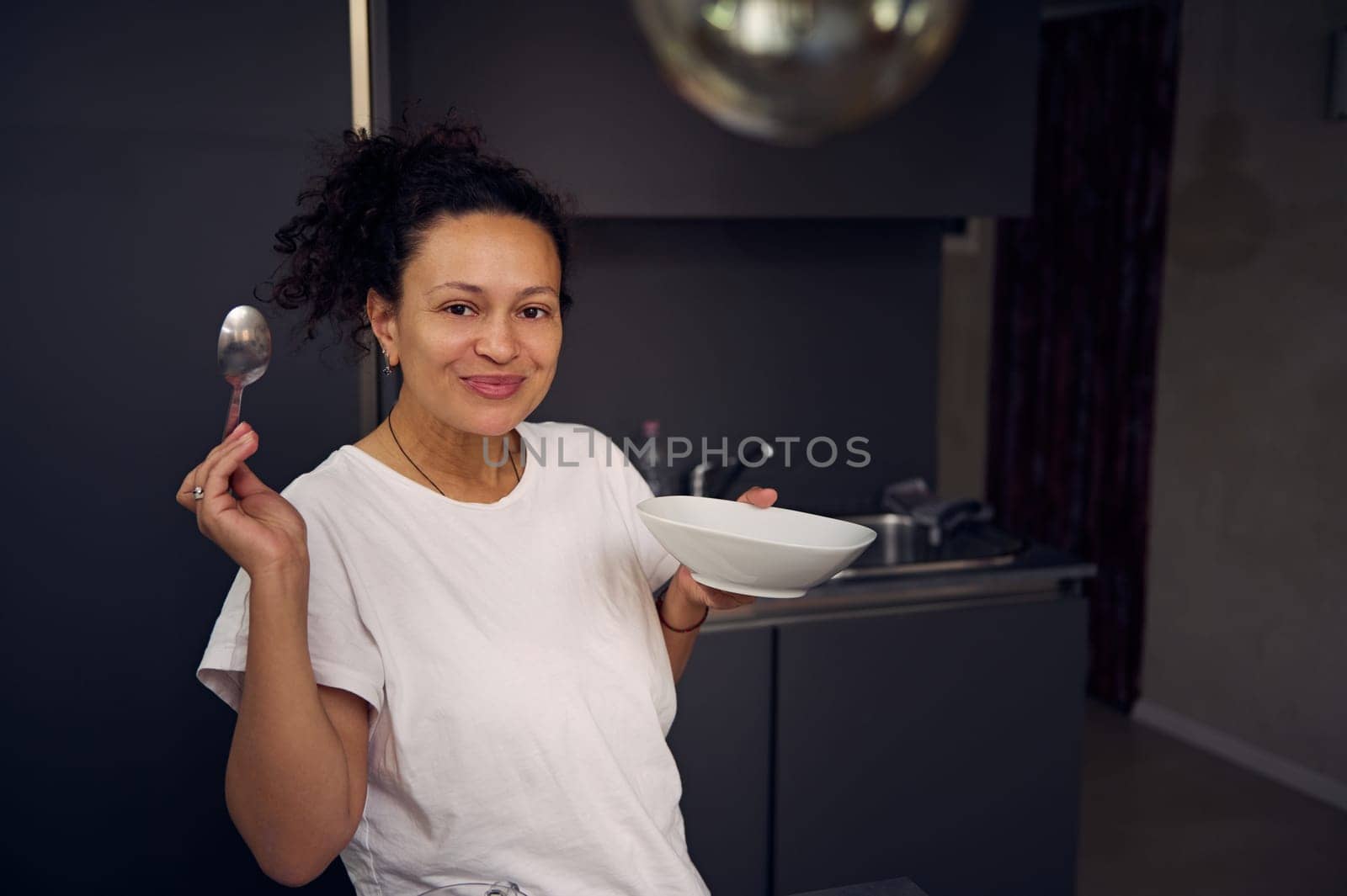 Beautiful curly haired brunette, young woman in pajamas, standing at home kitchen interior, smiling looking at camera, enjoying her healthy oat flakes for breakfast in the morning. People. Lifestyle