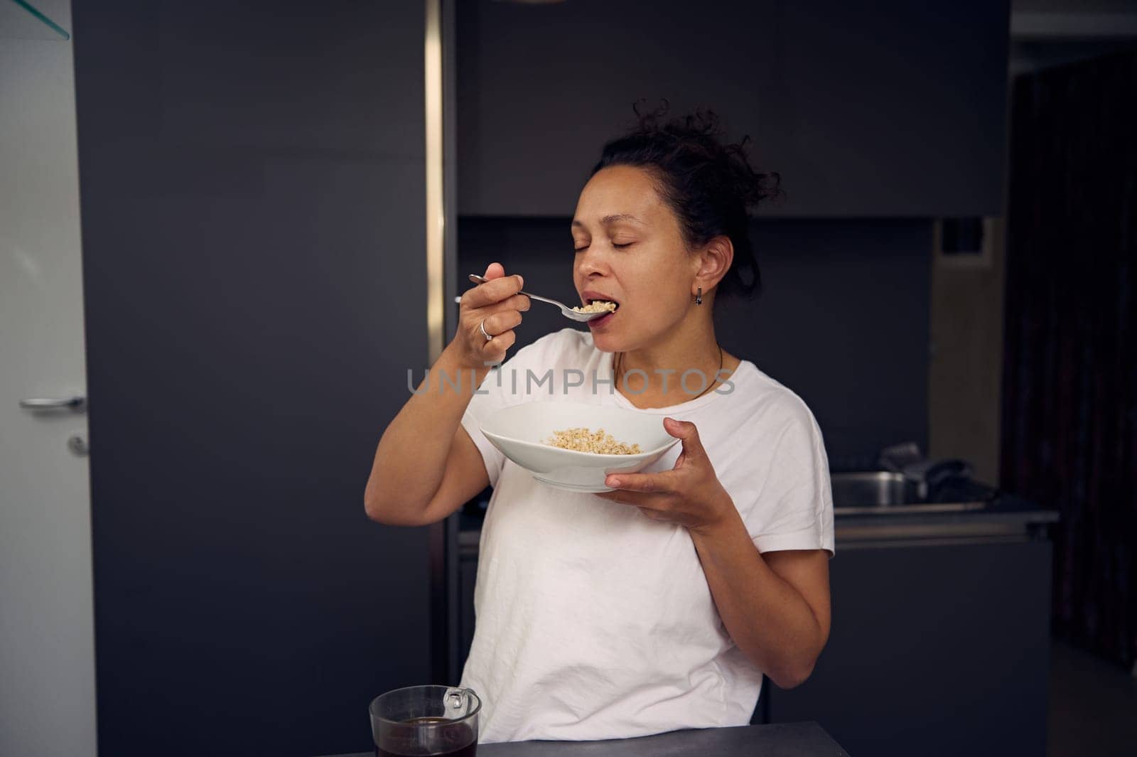 Happy Latina African American woman in pajamas, holding a muesli bowl in the home kitchen. Healthy lifestyle. People. Diet. Food consumerism.