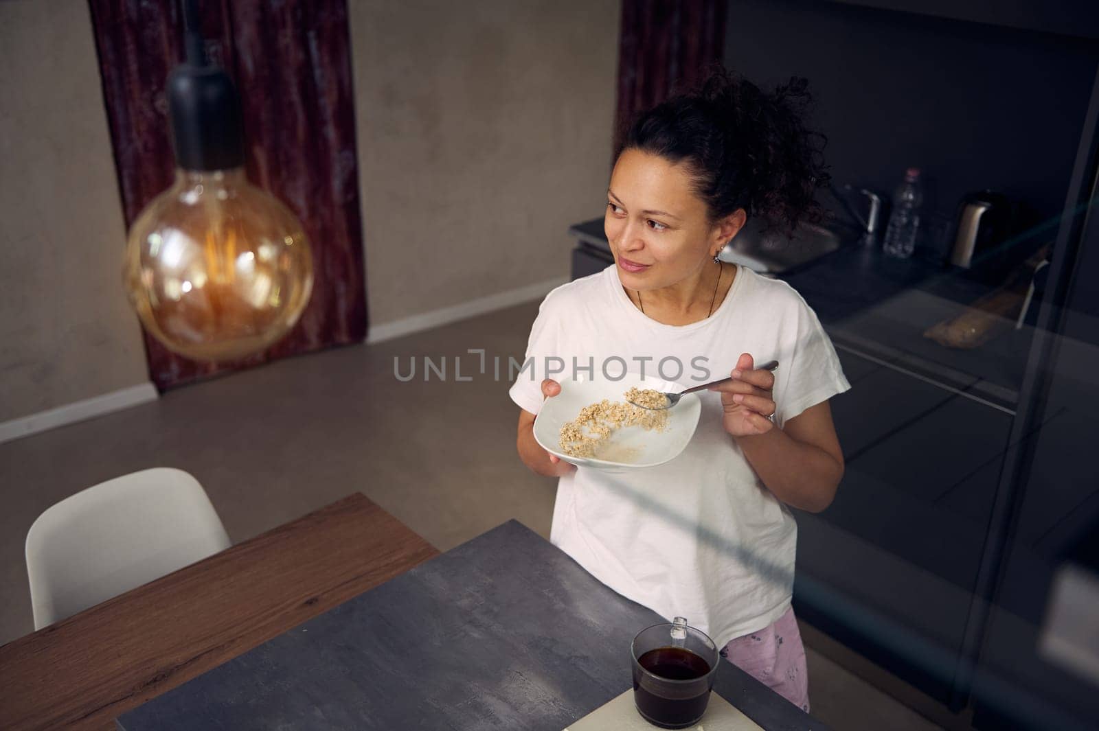 View from above of a charming confident woman at home, smiling looking aside, standing at kitchen island with a white muesli bowl in her hands, enjoying healthy oats for breakfast in the morning. Food