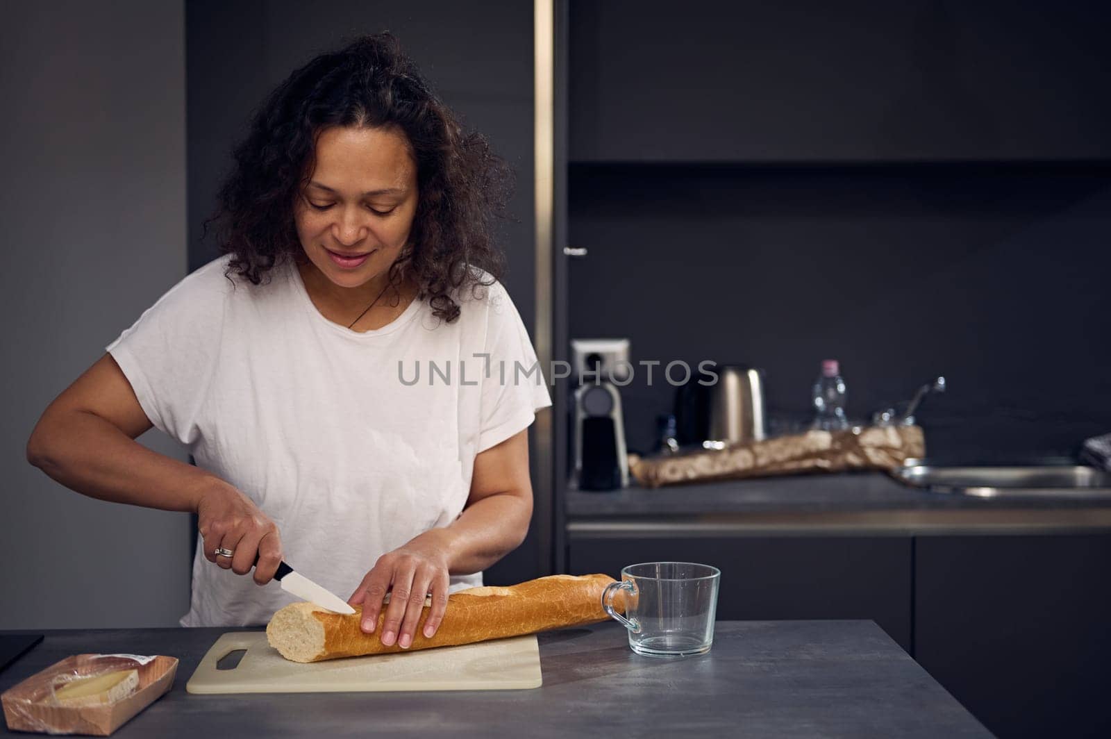 Pretty woman in white t-shirt, standing at kitchen counter, holding a kitchen knife nd cutting bread on the cutting board, preparing breakfast in the home kitchen in the morning