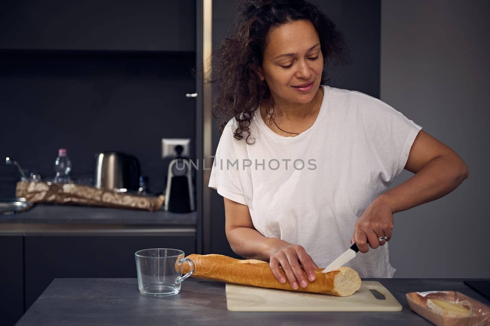 Mixed race beautiful woman in white t-shirt, standing at kitchen counter, holding knife and slicing a loaf of whole grain bread on cutting board, preparing breakfast in the home kitchen in the morning