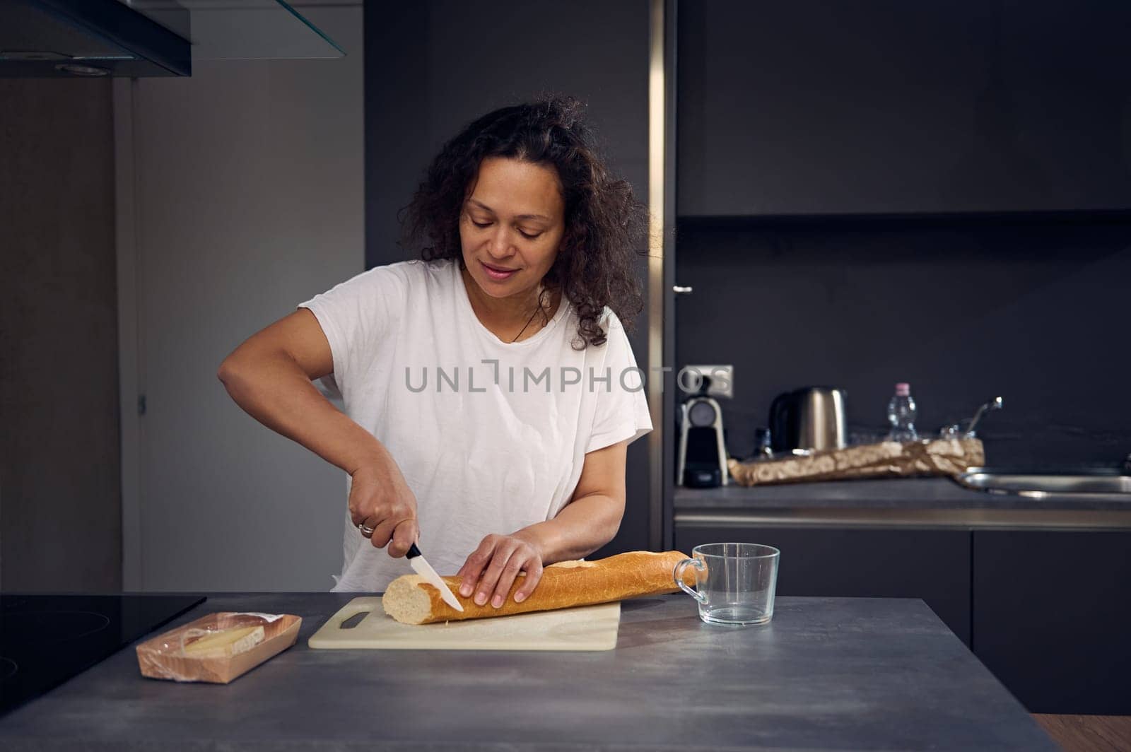 Mixed race beautiful woman in white t-shirt, standing at kitchen counter, holding knife and slicing a loaf of French baguette on a cutting board, preparing breakfast in the home kitchen in the morning