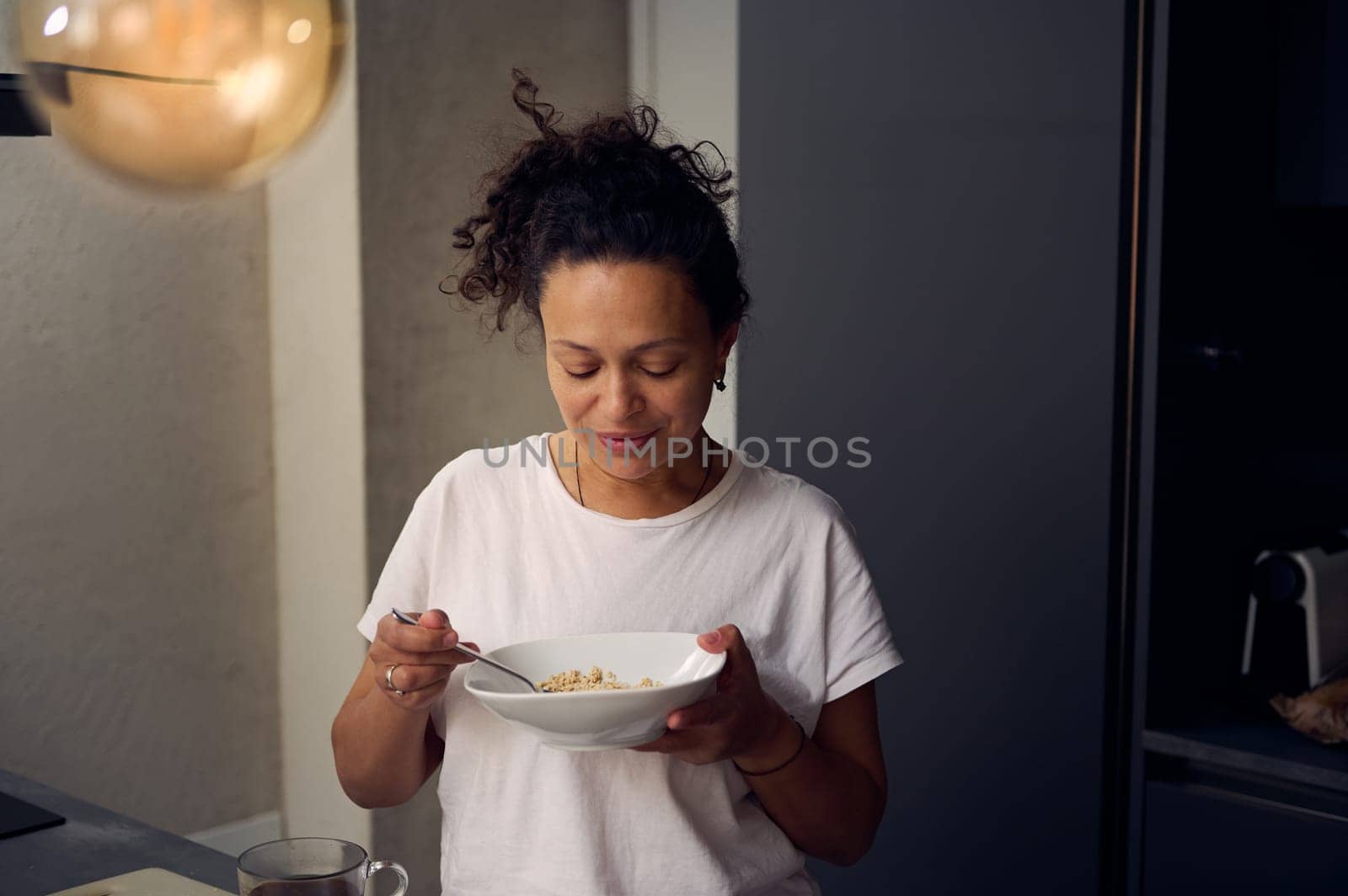 Young curly haired pretty woman in white t-shirt, holding a bowl of muesli, standing at kitchen counter at home. People. Food consumerism. Diet and slimming concept. Healthy lifestyle