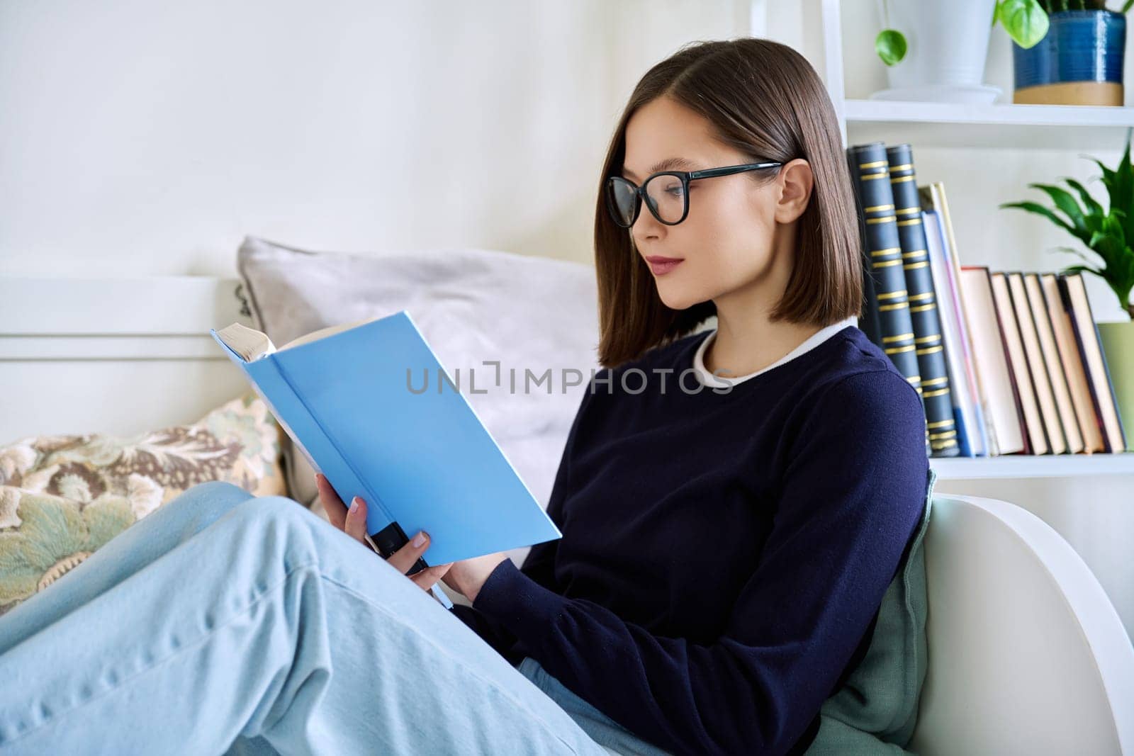 Young woman reading a book, sitting on the couch at home, engrossed in fiction. Literary hobby, education, lifestyle, youth concept