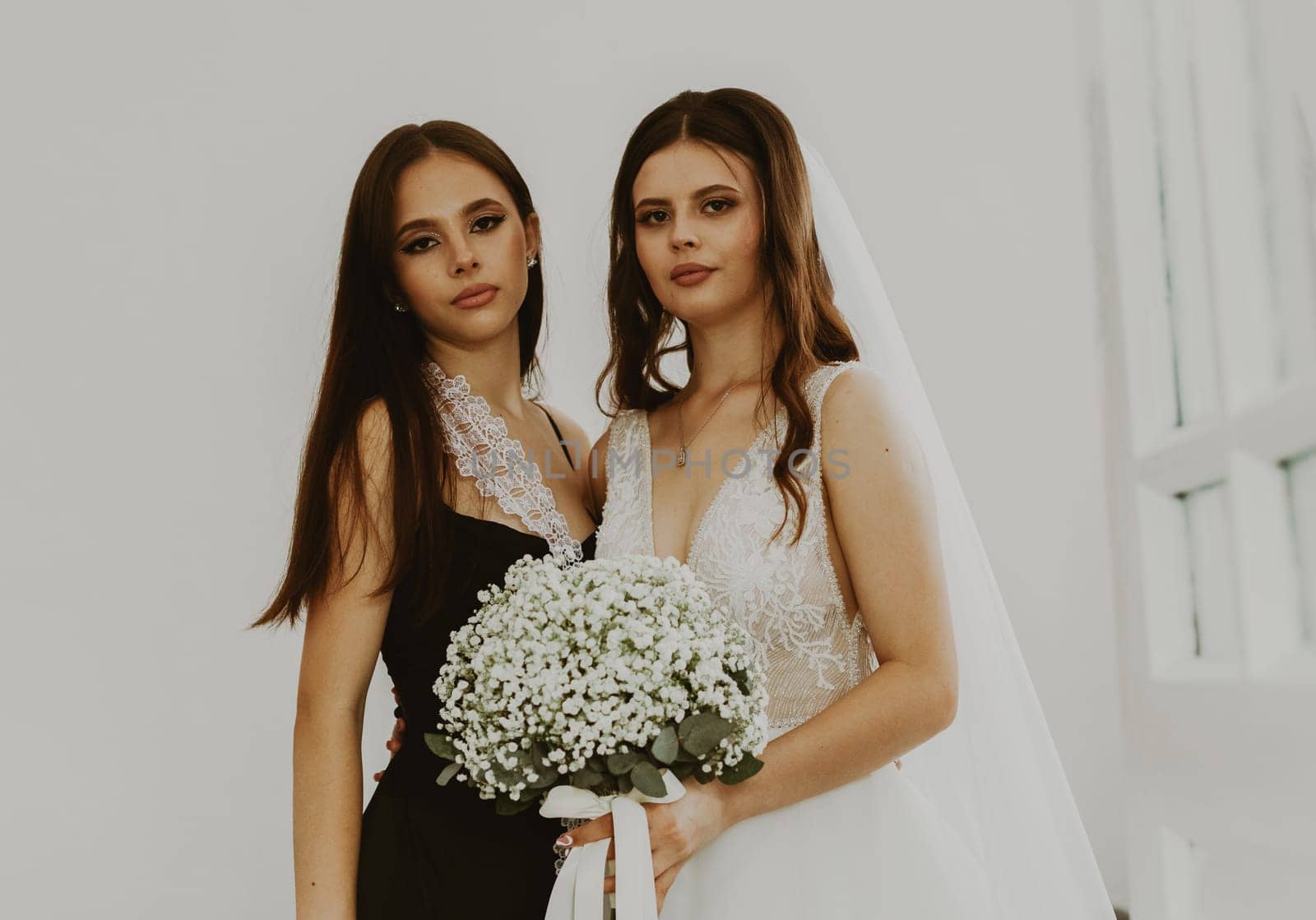 Portrait of a beautiful young Caucasian bride in a white dress with a wedding bouquet of white boutonnieres stands in an embrace with her bridesmaid, side view, close-up.