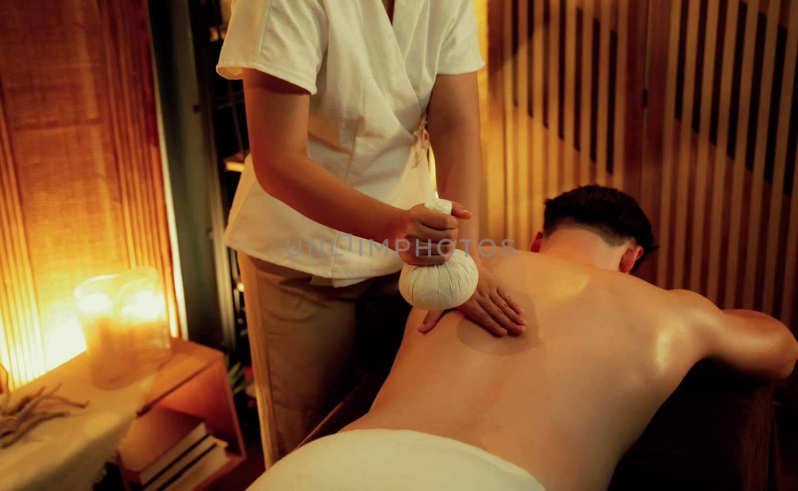 Hot herbal ball spa massage body treatment, masseur gently compresses herb bag on man body. Tranquil and serenity of aromatherapy recreation in warm lighting of candles at spa salon. Quiescent