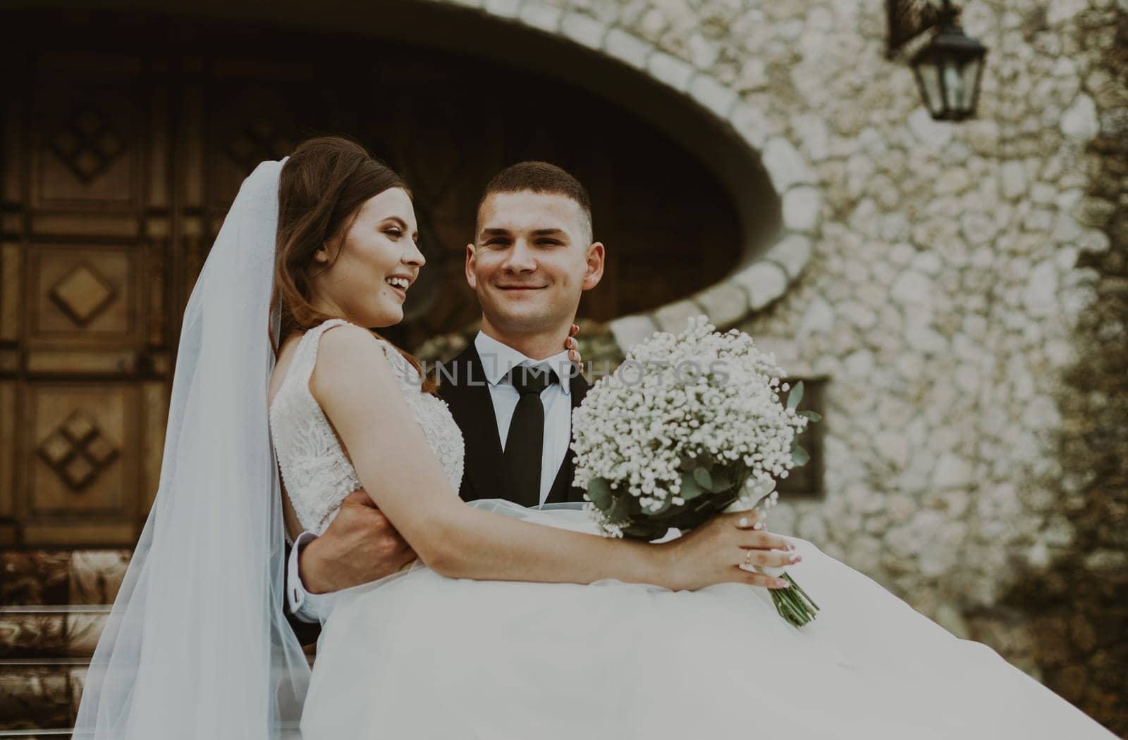 Portrait of one young beautiful stylish Caucasian happy and loving groom who holds his bride in his arms on a rainy cloudy day, close-up side view. Wedding traditions concept.