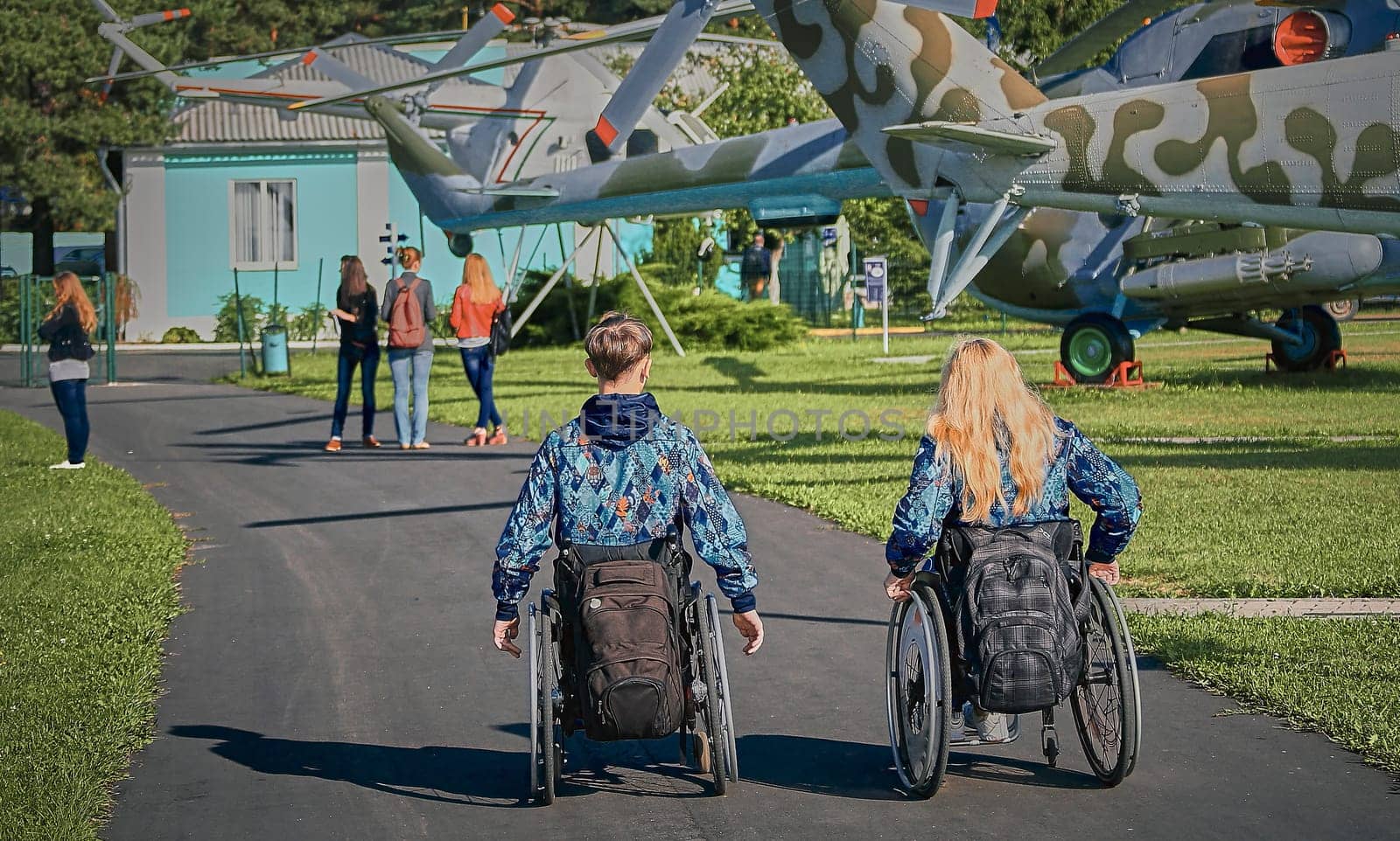 Two individuals, a man and a woman, both in wheelchairs, are exploring an outdoor museum featuring retired military helicopters on a sunny day, showcasing accessibility and inclusion.