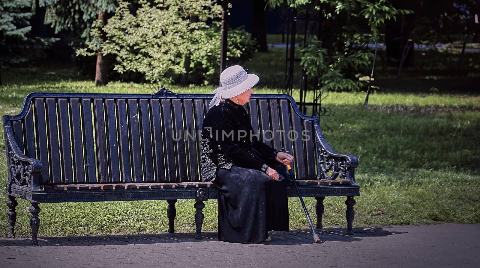 An elderly woman wearing a black dress and white hat sits on a park bench, surrounded by lush trees. The serene expression on her face suggests that she is enjoying the peace and quiet of the park.