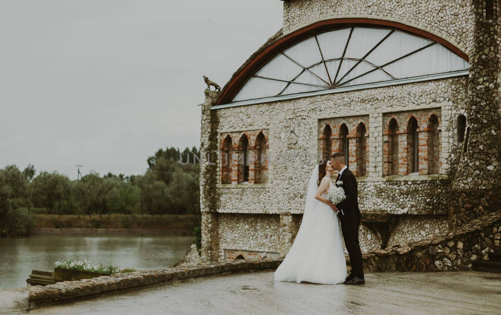 Portrait of one young beautiful stylish Caucasian woman kissing the bride and groom standing sideways with a bouquet of boutonnieres on a rainy cloudy day against the backdrop of an old castle with a lake, side view close-up with depth of field.